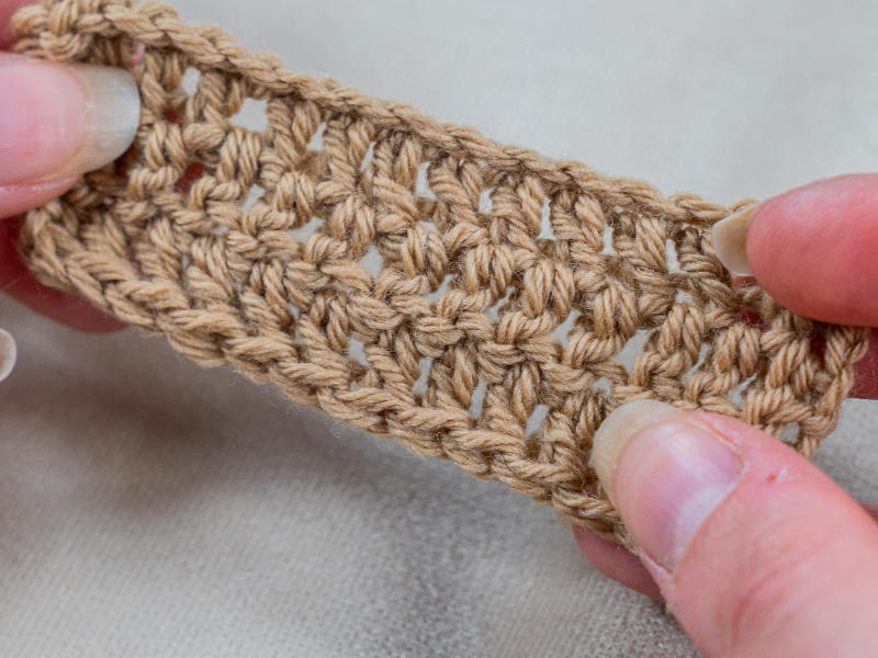 Gros plan de mains tenant un petit morceau rectangulaire de tissu beige demi bride au crochet, montrant des points détaillés sur un fond neutre.