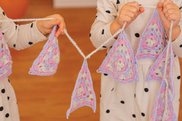 Deux enfants portant des robes blanches à pois noirs tiennent une chaîne de triangles de chignon au crochet de couleur pastel, parfaits pour ceux qui apprennent le point de crochet pour débutants. L'arrière-plan est légèrement flou.