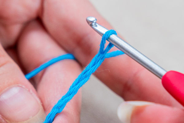 Gros plan sur des mains tenant un crochet argenté avec une poignée rouge, travaillant habilement un fil bleu pour former un point de maille en l'air au crochet.