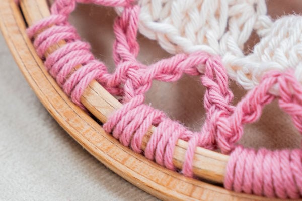 Gros plan sur un fil rose et blanc tissé de manière complexe autour d'un cercle à broder en bois, formant un motif décoratif inspiré des mandalas au crochet. L'arrière-plan est un tissu doux et neutre.