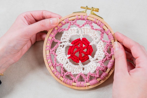 Deux mains tiennent un cercle à broder en bois avec une fleur au crochet inspirée des mandalas au crochet. La fleur a un centre rouge, un milieu blanc et des pétales extérieurs roses. Le cercle repose sur une surface de couleur claire.