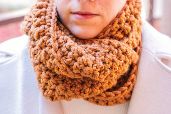 Une personne portant une grosse écharpe brune tricotée à la main avec du point de crochet pour débutants autour du cou et un manteau de couleur claire, vue du nez vers le bas.