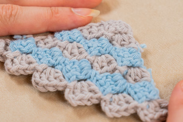 Gros plan des mains tenant une pièce de crochet c2c présentant un motif en damier avec une alternance de fils bleu clair et gris sur une surface beige.