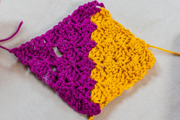 Un petit échantillon de c2c au crochet avec deux couleurs, le violet et le jaune, chaque couleur formant des sections distinctes. Les points créent un motif texturé, en blocs, sur un fond de tissu clair.