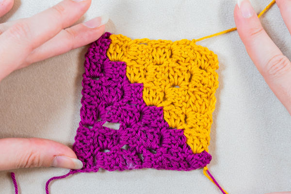 Gros plan de deux mains tenant un carré c2c au crochet, présentant un motif de coin à coin avec du fil violet à gauche et du fil jaune à droite.
