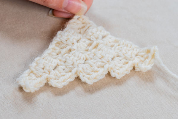 Une main tient un petit morceau triangulaire de crochet blanc c2c avec un motif texturé et festonné, reposant sur une surface de couleur claire.