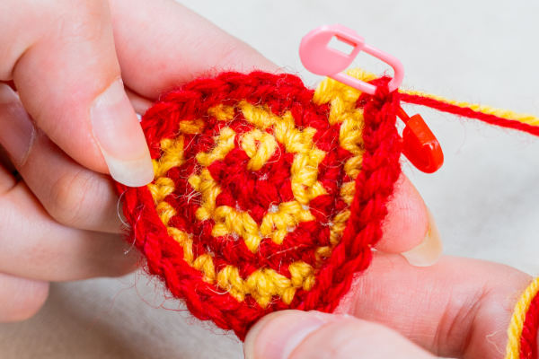 Gros plan des mains tenant une petite pièce circulaire crochetée avec des spirales rouges et jaunes, à l'aide d'un crochet et d'un marqueur de point rose - parfait pour toute personne apprenant à crocheter en spirale.