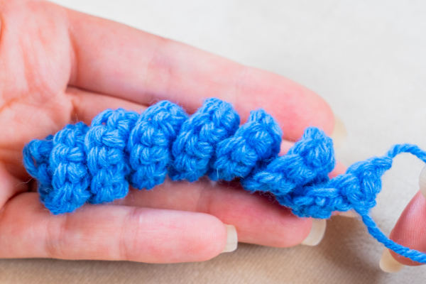 Une main tient une petite spirale bleue en fil crocheté serré, mettant en valeur la technique du crocheteur en spirale avec son motif texturé et ses points individuels.