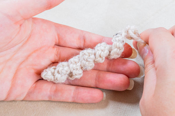 Gros plan d'une main tenant un petit morceau de fil crocheté blanc cassé, tandis qu'une autre main travaille selon la technique du crocheteur en spirale. Les points de crochet en spirale texturés sont clairement visibles.