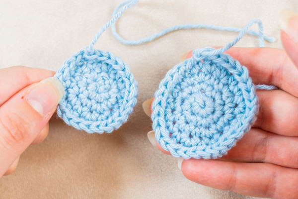 Deux mains tenant des cercles crochetés bleu clair avec des queues de fil attachées, démontrant la progression d'une technique de crochetage en spirale sur une surface neutre.