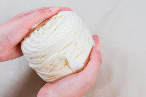 Gros plan de deux mains tenant délicatement une pelote ronde de fil trapilho XL pas cher ou des bandes de tissu sur un fond uni de couleur claire.
