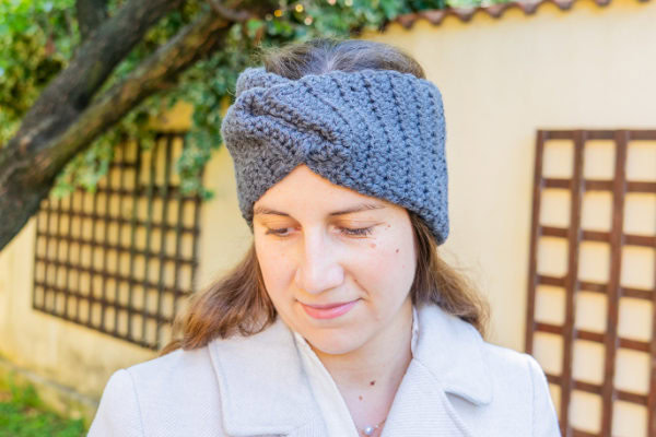 Une femme portant un bandeau gris texturé au crochet - parfait pour ceux qui souhaitent apprendre le crochet - se tient à l'extérieur, regardant vers le bas avec un léger sourire. Elle est vêtue d'un manteau clair, entourée de verdure et d'un mur crème.