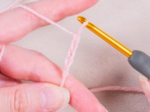 Gros plan sur des mains tenant un fil rose clair et un crochet doré, formant une chaîne au crochet sur un fond beige - parfait pour ceux qui souhaitent apprendre le crochet.