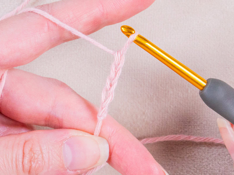 Gros plan sur des mains tenant un fil rose clair et un crochet doré, formant une chaîne au crochet sur un fond beige - parfait pour ceux qui souhaitent apprendre le crochet.