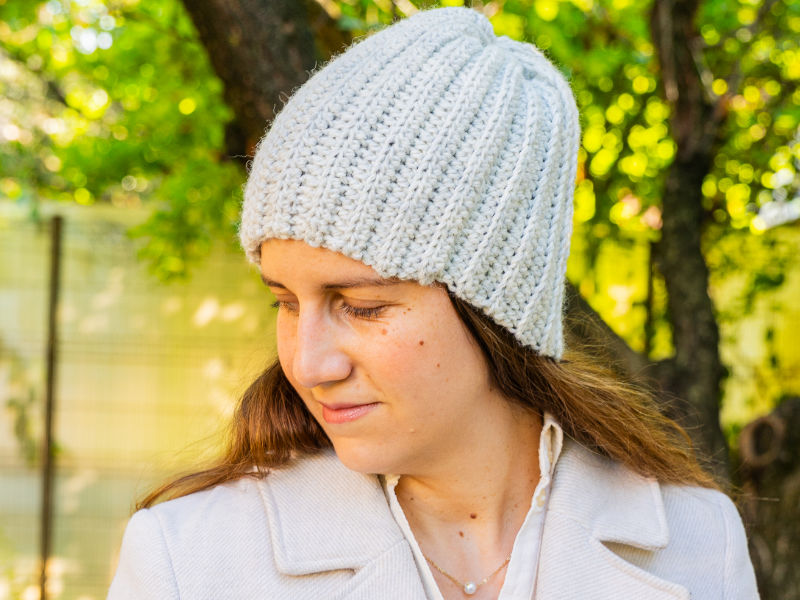 Une femme portant un bonnet en tricot de couleur claire et un manteau crème se tient à l'extérieur, près d'arbres, regardant vers le bas et souriant doucement, comme si elle était fière de crocheter un bonnet. L'arrière-plan est vert et ensoleillé.