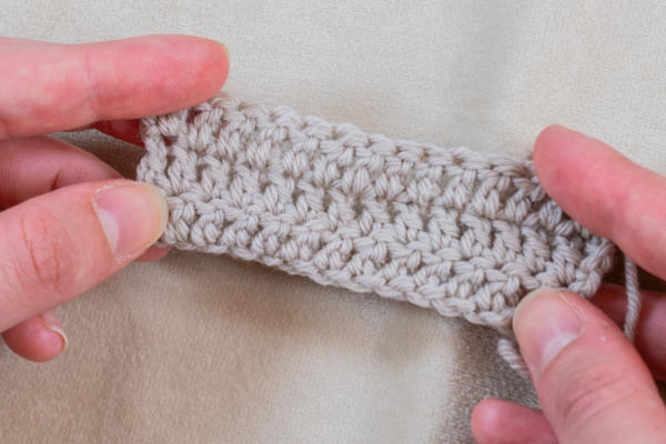 Deux mains tiennent un petit morceau rectangulaire de tissu crocheté beige clair, mettant en valeur quelques rangs de fil cousu serré. Parfait pour un projet de crocheter un coeur, le fond doux et neutre met en valeur chaque détail délicat.