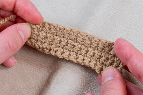 Gros plan de deux mains tenant un petit morceau rectangulaire d'ouvrage au crochet de couleur havane sur un fond de tissu beige. La technique "crocheter un cœur" est mise en valeur par les points serrés et régulièrement espacés.
