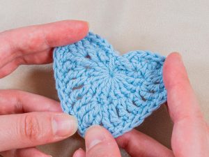 Deux mains tiennent délicatement un petit cœur bleu clair crocheté sur un fond de tissu beige, illustrant l'art délicat de crocheter un cœur.