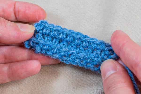Deux mains tiennent un petit morceau de tissu bleu au crochet, montrant les points texturés sur un fond beige clair - une inspiration parfaite pour tout projet de crocheter un cœur.