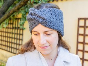 Une femme aux cheveux bruns porte un épais bandeau gris tricoté au crochet et un manteau de couleur claire, debout en plein air près d'un mur et d'un arbre. Elle regarde vers le bas avec une expression calme.