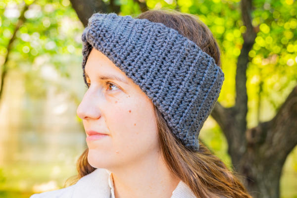 Une femme aux cheveux châtain clair porte un épais bandeau gris au crochet en plein air. Elle regarde vers la gauche, avec des arbres verts et feuillus en arrière-plan.