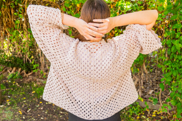 Une personne se tient à l'extérieur, les mains derrière la tête, vêtue d'un pull-over au crochet ample et de couleur claire - une inspiration parfaite si vous vous demandez comment faire un gilet au crochet facile. Le feuillage vert et la lumière du soleil remplissent l'arrière-plan.
