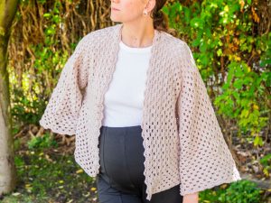 Une personne portant un cardigan beige clair, crocheté et ouvert sur le devant - une inspiration parfaite si vous cherchez comment faire un gilet au crochet facile - se tient à l'extérieur devant un feuillage vert. Le cardigan a des manches larges et un motif texturé et aéré.