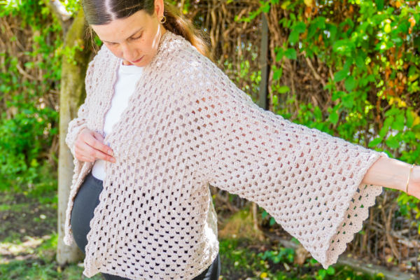 Une femme portant un gilet au crochet beige clair - une inspiration parfaite si vous voulez comment faire un gilet au crochet facile - écarte un bras pour montrer le détail de la manche alors qu'elle se tient à l'extérieur dans un jardin verdoyant.