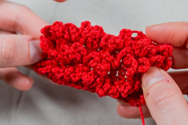 Deux mains tiennent un morceau de crochet rouge vif, texturé, comportant le point d'astrakan au crochet, avec un motif de volutes et de couches sur un fond neutre.