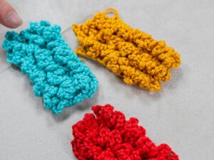 Trois rectangles texturés, crochetés à la main en fil bleu, jaune et rouge - réalisés au point d'astrakan au crochet - reposent sur une surface gris clair. Le doigt d'une personne est visible près du morceau bleu dans le coin supérieur gauche.