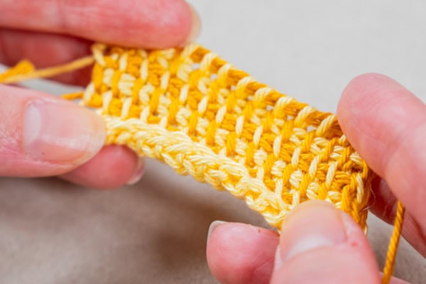 Gros plan de mains tenant un petit morceau de tissu jaune crocheté, présentant des points de crochet détaillés selon la technique du crochet tunisien.