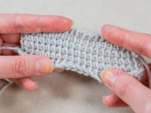 Gros plan de deux mains tenant un morceau de tissu tunisien gris clair au crochet, montrant clairement la texture et les points, sur un fond neutre.