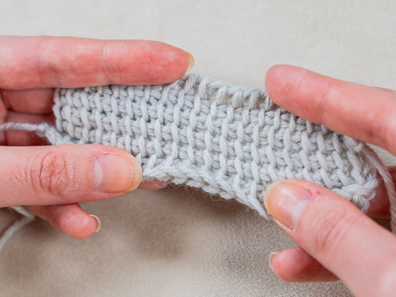 Gros plan de deux mains tenant un morceau de tissu tunisien gris clair au crochet, montrant clairement la texture et les points, sur un fond neutre.