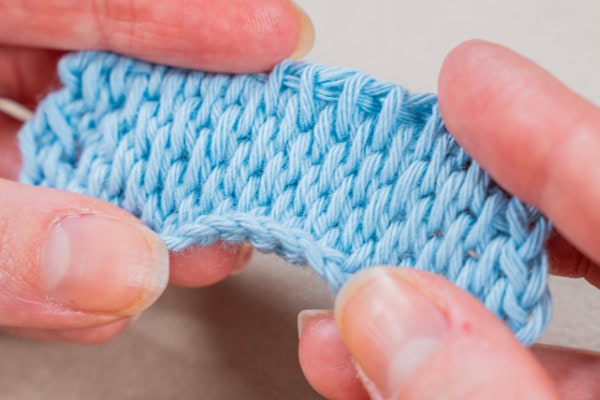Gros plan de deux mains tenant et étirant légèrement un morceau de tissu tunisien bleu clair au crochet, montrant la texture et le motif des points.