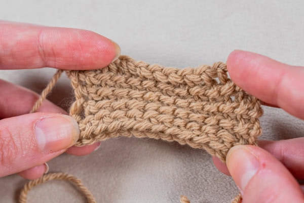 Gros plan de deux mains tenant un petit morceau rectangulaire de tissu tunisien beige au crochet, mettant en évidence la texture et les points détaillés du fil.