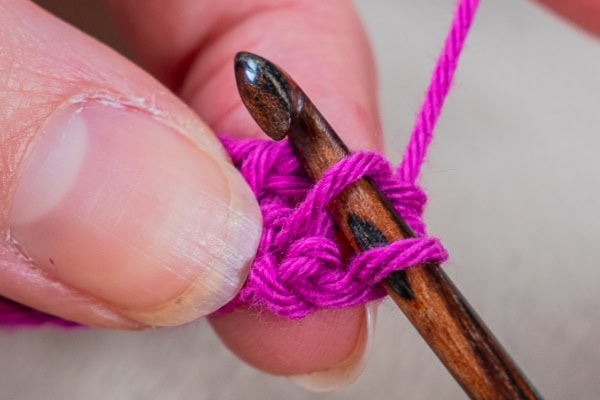 Gros plan d'une main tenant un crochet travaillant avec un fil violet vif, montrant le crochet inséré dans une maille et le fil enroulé autour, selon la technique du crochet tunisien.