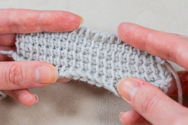 Gros plan de deux mains tenant un morceau de tissu tunisien gris clair au crochet, montrant les détails texturés des points uniques. L'arrière-plan est une surface douce et neutre.