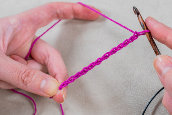 Gros plan de mains tenant un crochet et travaillant avec un fil rose vif, créant une chaîne de points de crochet tunisiens sur un fond neutre.