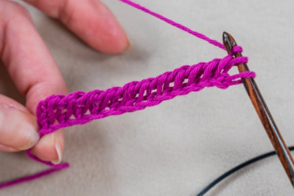 Gros plan d'une main tenant un fil rose vif et crochetant à la tunisienne avec un crochet en bois, montrant plusieurs points terminés sur un fond neutre.