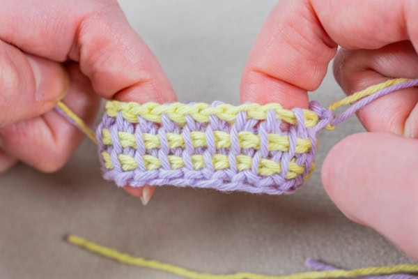 Gros plan des mains tenant un petit morceau de crochet tunisien avec des rangées alternées de fils violet clair et jaune, montrant le motif de point en détail.