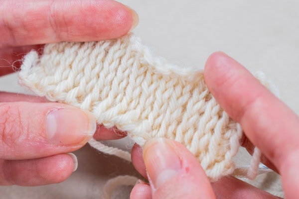 Gros plan de deux mains tenant un petit morceau de tissu tunisien blanc au crochet, mettant en évidence la texture détaillée et les points du fil.