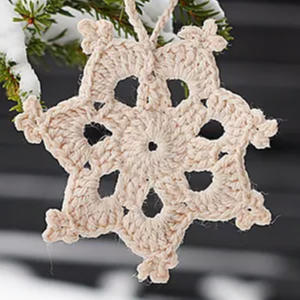 Un flocon de neige beige crocheté est suspendu à une branche de conifère enneigée, avec un arrière-plan sombre et flou derrière lui.