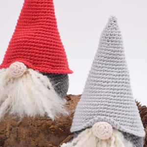 Deux poupées gnomes au crochet avec de longues barbes blanches et des chapeaux pointus, l'une rouge et l'autre gris clair, positionnées sur une surface brune sur un fond clair.