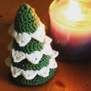 Une petite décoration de Noël tricotée, verte et blanche, est posée sur une surface en bois à côté d'une bougie allumée dans un bocal en verre.