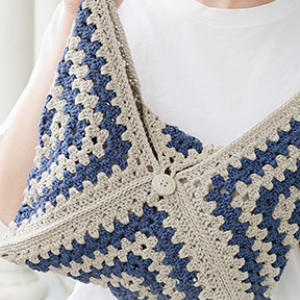 Une personne tenant un sac crocheté à la main avec des motifs granny square bleus et beiges, plié et fermé par un bouton, vêtue d'une chemise blanche.