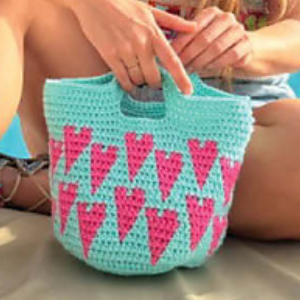 Une personne tient un sac crocheté bleu clair décoré de rangées de cœurs roses. Ses jambes sont visibles à l'arrière-plan et elle est assise, le sac reposant sur ses genoux - une inspiration parfaite pour un modèle sac crochet gratuit à télécharger.