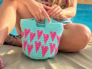 Une personne est assise au bord d'une piscine et tient un sac crocheté de couleur aqua avec des motifs de cœurs roses - un modèle de sac crochet gratuit à télécharger. Ses jambes sont croisées et ses mains reposent délicatement sur le sac posé sur ses genoux.