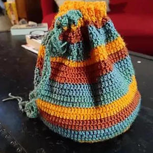 Un sac à cordon coloré au crochet avec des rayures horizontales en sarcelle, orange, jaune et rouge est posé sur une table sombre avec un canapé rouge en arrière-plan - parfait pour ceux qui recherchent un modèle sac crochet gratuit à télécharger.