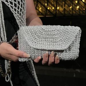 Une personne tient une pochette au crochet argentée avec explication gratuite, dotée d'une fermeture à rabat, alors qu'elle porte un ensemble noir avec un gilet en crochet assorti et une ceinture en chaîne dorée.