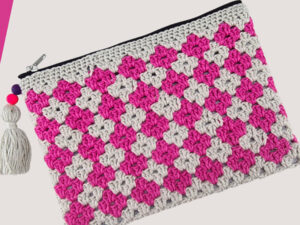 Une pochette au crochet à fermeture éclair avec un motif à carreaux roses et blancs, avec un pompon blanc et des perles colorées attachées à la fermeture éclair - une charmante pochette au crochet avec explication gratuite pour vos objets essentiels.
